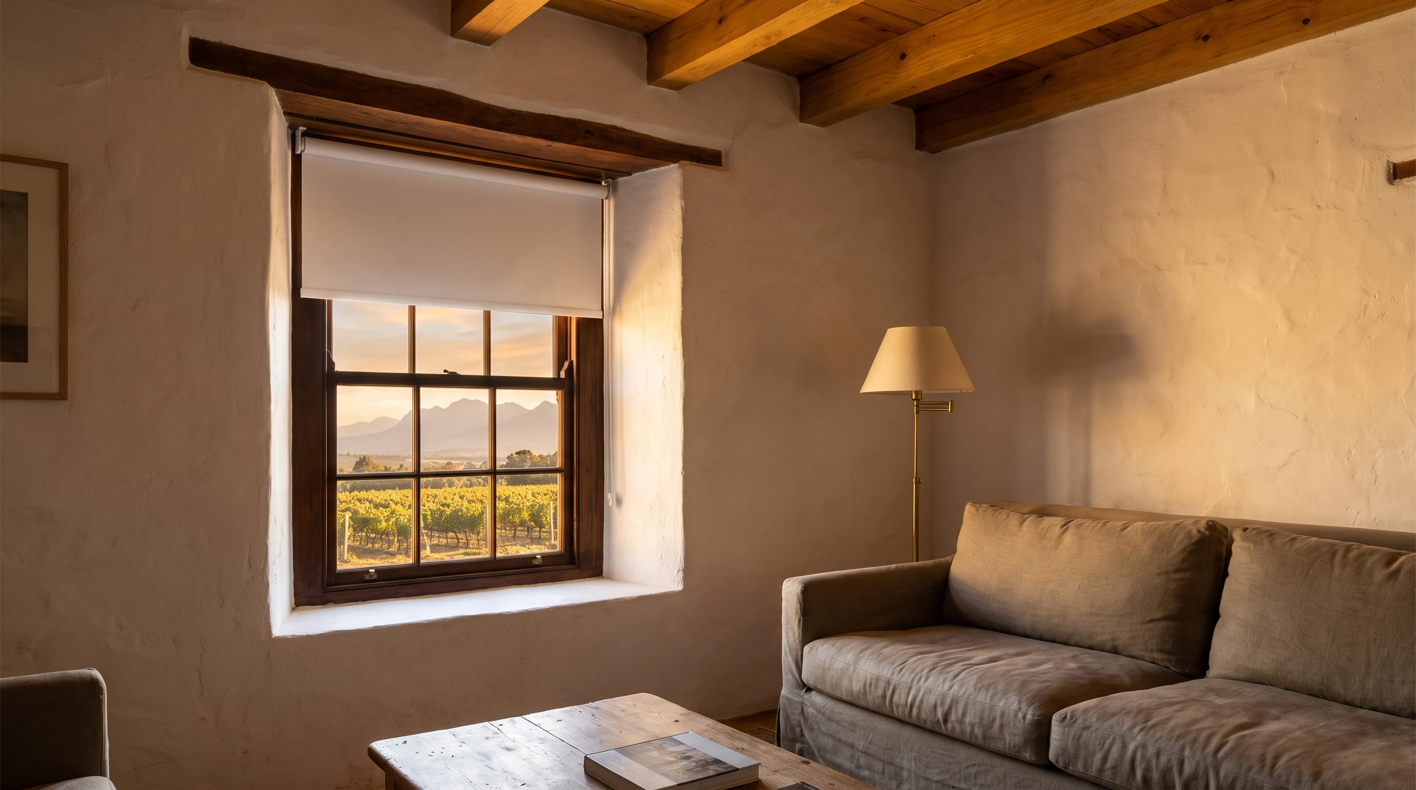 Living room in a Cape Dutch farmhouse near Groot Drakenstein with a smooth blockout roller blind half-lowered against a deep-set window, vineyard rows and the Groot Drakenstein mountains visible outside